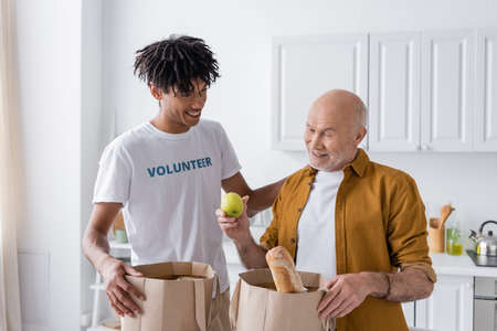 Smiling african american volunteer standing near pensioner and packages with food in kitchenの写真素材
