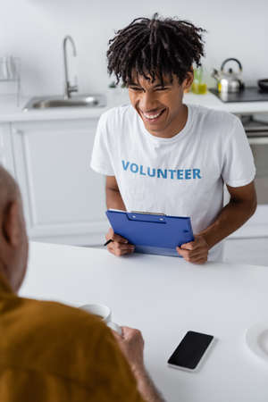 Cheerful african american volunteer with clipboard looking at blurred senior man with tea near...