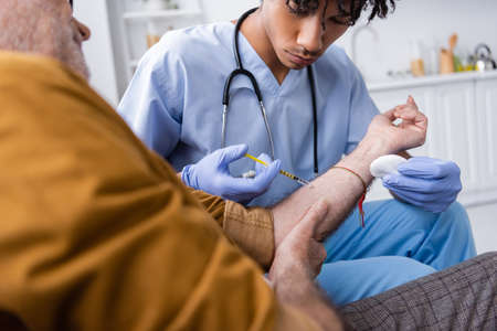 African american nurse in latex gloves and uniform doing injection to elderly patient at homeの写真素材