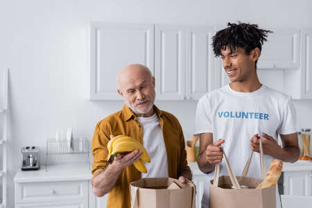 Smiling senior man holding bananas near bags and african american volunteer in kitchen