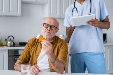Senior man holding pills near water and african american nurse using digital tablet in kitchen