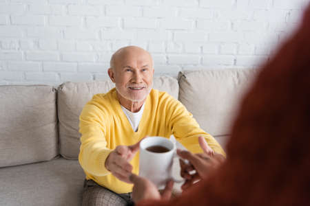 Blurred african american man holding cup of tea near smiling granddad on couch
