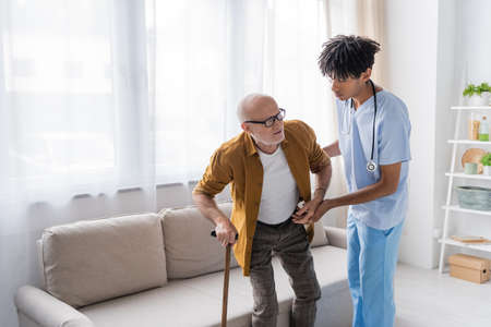 African american nurse in uniform helping elderly man with walking cane at home