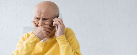 Upset elderly man talking on smartphone in living room, banner