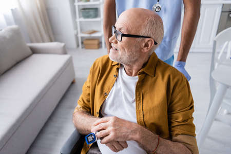 Smiling senior man with pulse oximeter looking away near african american nurse at homeの写真素材