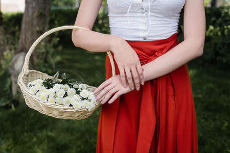 Cropped view of woman in blouse and skirt holding wicker basket with flowers in parkの写真素材