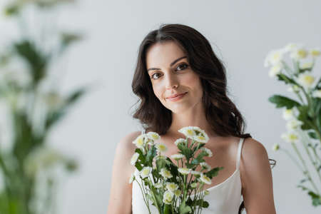 Smiling woman in white top looking at camera near blurred flowers isolated on greyの写真素材