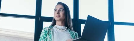 Low angle view of queer designer holding laptop in office, bannerの写真素材