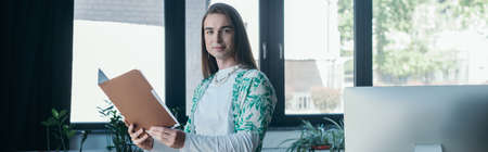 Young queer designer holding paper folder and looking at camera near computer in office, banner