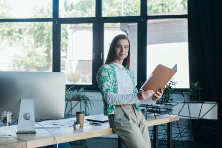 Smiling queer designer holding paper folder and looking at camera near working table in officeの写真素材