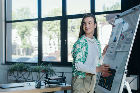 Smiling queer designer standing near graphs on flip chart in officeの写真素材