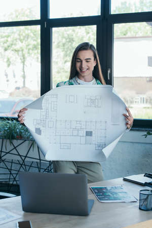 Smiling queer designer holding blueprint during video call on laptop in creative agency