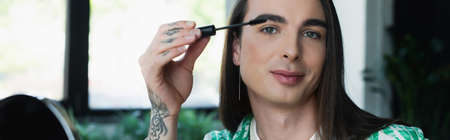 Nonbinary designer holding mascara brush and looking at camera in office, bannerの写真素材