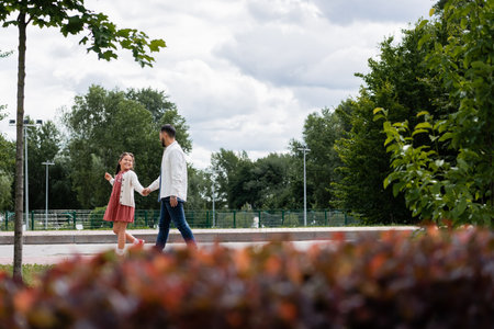 Smiling asian girl holding hand of father while walking in summer parkの写真素材