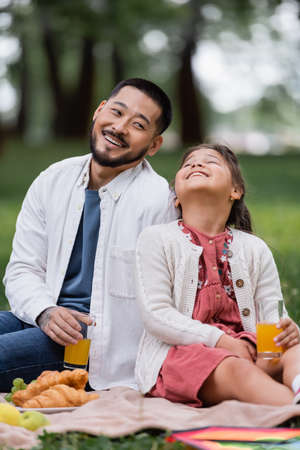 Positive asian family holding orange juice near croissants on blanket in parkの写真素材