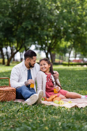 Asian father hugging daughter with orange juice near food on blanket in summer parkの写真素材