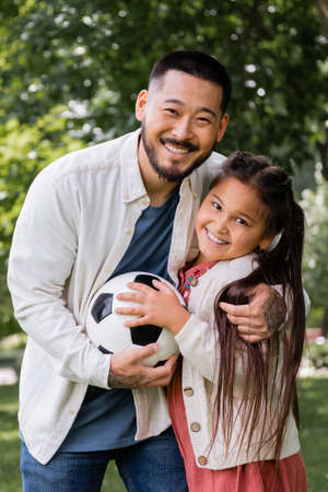 Positive asian dad and girl holding soccer ball and looking at camera in parkの写真素材