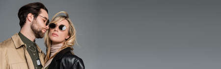 stylish bearded man in sunglasses looking at blonde woman in black leather jacket isolated on grey,...