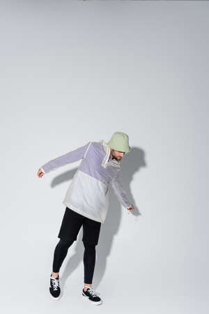 full length of young man in stylish sportive clothing and panama hat posing on grey