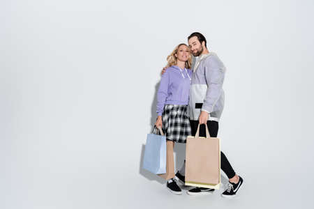 full length of happy man and blonde woman in tartan skirt holding shopping bags on grey
