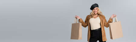 happy woman in stylish leather beret and beige blazer holding shopping bags isolated on grey, bannerの写真素材