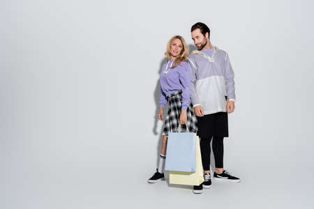 full length of happy woman in plaid skirt holding shopping bags near bearded man on grey