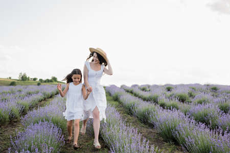 cheerful mother and daughter in white dresses holding hands while running in meadowの写真素材