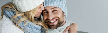 blonde woman in knitted hat hugging smiling boyfriend in winter outfit isolated on grey, banner