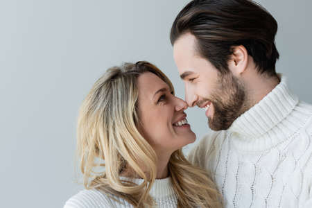 portrait of cheerful couple in knitted white sweaters smiling while looking at each other isolated on greyの写真素材