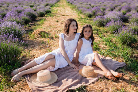 woman and girl in white dresses sitting on blanket in meadow near straw hatsの写真素材