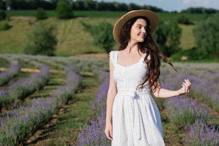 positive woman in white dress and straw hat looking away in meadow with blooming lavenderの写真素材