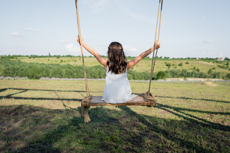 back view of child in summer dress riding swing in meadowの写真素材