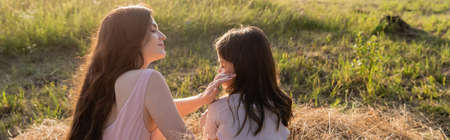 woman with long hair touching face of daughter while sitting in meadow, banner
