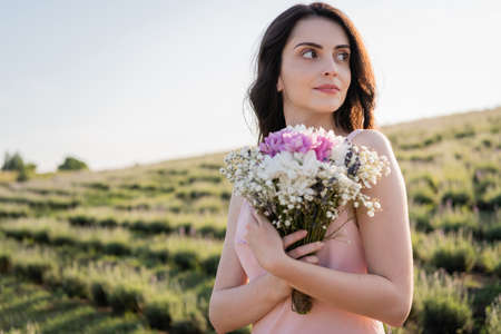 brunette woman with bouquet of fresh flowers looking away in countrysideの写真素材