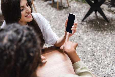 African american woman pointing with finger near friend with smartphone on cafe terraceの写真素材