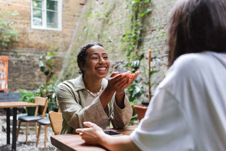 Cheerful african american woman talking to friend near smartphone in outdoor cafeの写真素材