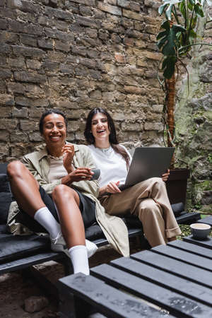 Cheerful multiethnic girlfriends with coffee and laptop looking at camera on terrace of cafeの写真素材