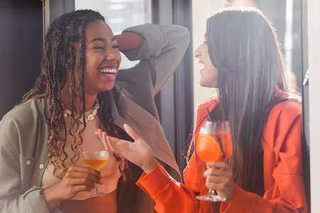 Positive multiethnic girlfriends holding cocktails and talking near window in cafeの写真素材