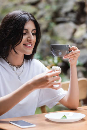 Positive brunette woman holding dessert and coffee near cellphone on terrace of cafe