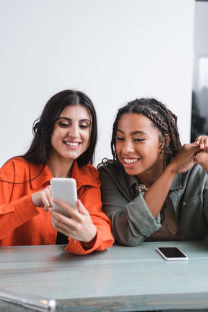 Cheerful african american woman looking at cellphone near friend in cafeの写真素材