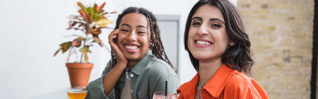 Cheerful woman looking at camera near african american friend and cocktails in cafe, banner