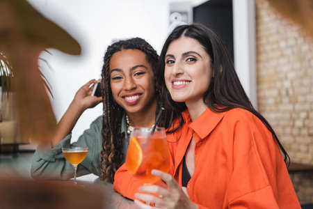 Smiling multiethnic girlfriends with smartphone looking at camera near cocktail in cafe