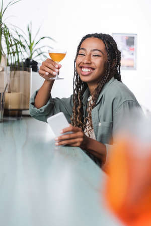 African american woman holding glass of cocktail and smartphone while smiling at camera in cafeの写真素材