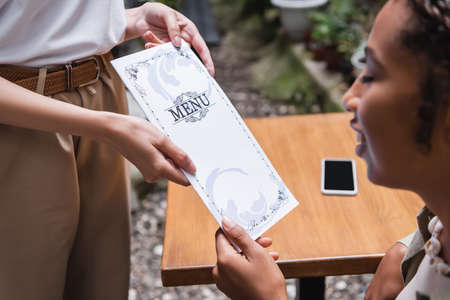 Blurred african american woman taking menu from waitress on terrace of cafeの写真素材