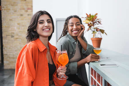 Smiling woman holding cocktail near blurred african american friend and smartphones in cafe