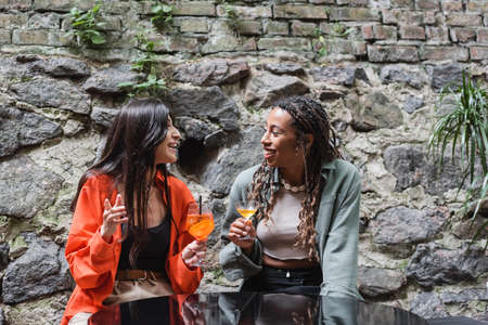Smiling interracial girlfriends with cocktails talking on terrace of cafeの写真素材