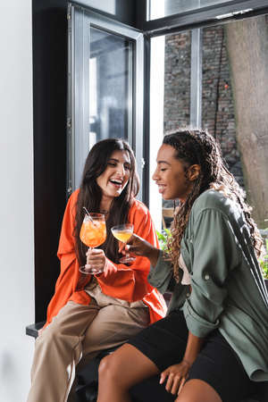 Positive interracial girlfriends holding cocktails on windowsill in cafeの写真素材