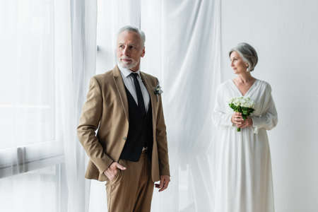 bearded middle aged man in suit posing with hand in pocket near blurred bride in white dress standing with bouquet near white curtainsの写真素材