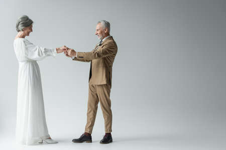 full length of cheerful middle aged man in suit and bride in white wedding dress holding hands on...