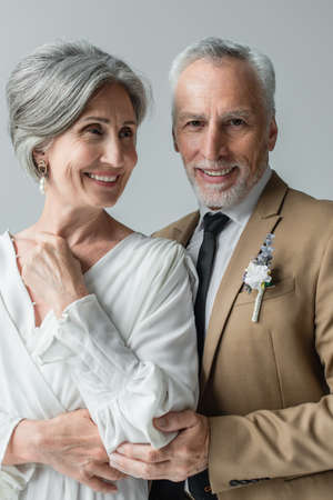 happy middle aged man in suit with floral boutonniere and woman in white wedding dress isolated on greyの写真素材